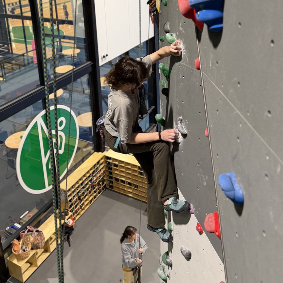 A young climber on top rope high on the grey slab wall at Beta Park at the Hub, belayed from the ground by another climber